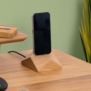 Op de afbeelding: Een houten telefoonstandaard met een geometrisch ontwerp. Een zwarte smartphone staat op de standaard, met een zwarte kabel die uit de basis komt. De standaard is gemaakt van lichtgekleurd hout en staat op een houten bureau.