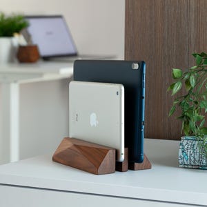 May include: A wooden tablet stand holding two tablets, one silver and one dark blue. The stand has a geometric design and is made of dark wood. The tablets are displayed on a white surface.