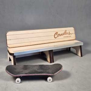 May include: A miniature wooden bench with the text "Cornelius Fingerboard" and a small skateboard. The bench has a light wood finish with silver accents. The skateboard has a black grip tape surface and white wheels.
