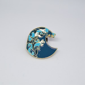 Könnte beinhalten: Eine blaue und weiße Emaille-Anstecknadel in Form einer Welle mit goldenen Akzenten.
