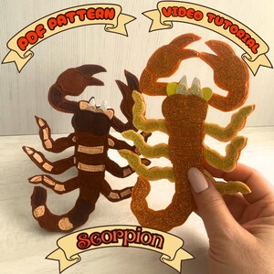 May include: Two felt scorpion crafts in shades of brown and orange. One is held by a hand. The image includes text that reads "PDF Pattern", "Video Tutorial", and "Scorpion".