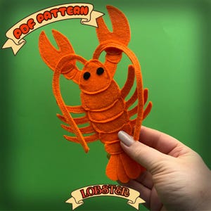 May include: A bright orange felt lobster craft with black eyes and detailed claws. The image includes the text "PDF PATTERN" and "LOBSTER" on banners. The lobster is held against a green background.