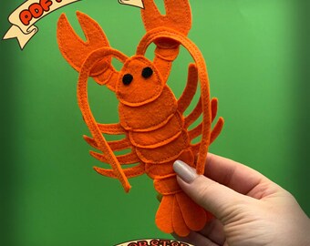 May include: An orange felt lobster craft with black eyes and detailed claws. The image includes the text "PDF PATTERN" and "LOBSTER" on banners. The lobster is held against a green background.