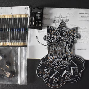 May include: An overhead shot of electronic components for a DIY project. Includes resistors, integrated circuits, a circuit board with a graphic design, and a bag of smaller parts. The circuit board has the text "Warfare".
