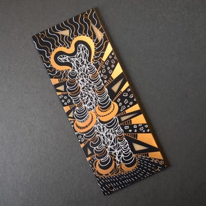 May include: A rectangular, black and gold decorative item with an abstract design. The design features white, black, and gold geometric shapes and patterns. The item has two small holes at the top for hanging.