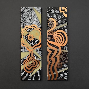 May include: Two abstract art bookmarks. One features a gold and black design with a flower-like shape and lightning bolts. The other has a copper and black design with swirling lines and star shapes. Both have a hole at the top.