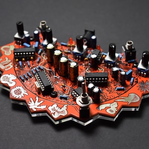 May include: A red and black circuit board with various electronic components, including resistors, capacitors, and potentiometers. The board is shaped like a star and has a white design with small drawings of animals. The text "THE KNUFFYI 17" is printed on the board.