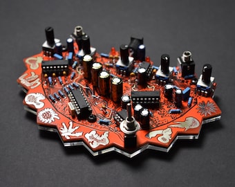 Puede incluir: Una placa de circuito rojo y negro con varios componentes electrónicos, incluyendo resistencias, condensadores y potenciómetros. La placa tiene forma de estrella y un diseño blanco con pequeños dibujos de animales. El texto "THE KNUFFYI 17" está impreso en la placa.