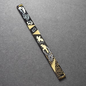 May include: A black and gold decorative bracelet with a geometric design. The bracelet features alternating black and gold sections, with white abstract shapes and patterns. The bracelet has a small hole at one end.