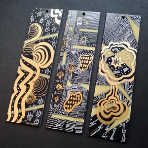 Può includere: Tre segnalibri rettangolari con intricati motivi grafici in nero, bianco e oro. Ogni segnalibro presenta un piccolo foro circolare nella parte superiore e inferiore. I disegni includono linee vorticose, forme geometriche e il testo ISN7S1S.