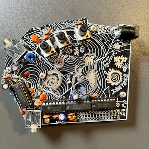 Könnte beinhalten: Eine schwarze Leiterplatte mit weißen dekorativen Linien und dem Text "FORT PROCESSOR ISN'TS". Sie verfügt über verschiedene elektronische Komponenten in Orange, Blau und Silber, darunter Knöpfe und Chips.