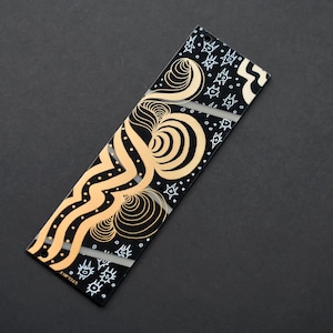 May include: A rectangular, black decorative item with an abstract design in gold and white. The design features swirling lines, dots, and geometric shapes. The item is set against a dark gray background.