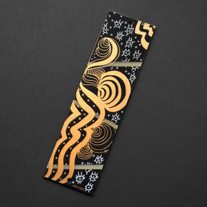 May include: A rectangular black bookmark with an abstract design in gold and white. The design features swirling lines, dots, and star-like shapes. The bookmark is a unique and artistic accessory for book lovers.