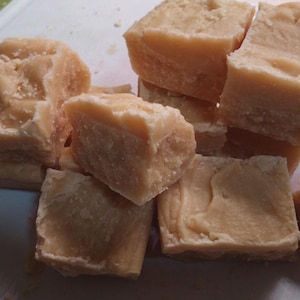 Könnte beinhalten: Eine Ansammlung hellbrauner Fudge-Quadrate, einige gestapelt, auf einem weißen Schneidebrett. Der Fudge hat eine leicht raue Textur und scheint frisch geschnitten zu sein. Die Farbe ist ein cremiges, helles Braun, was auf einen klassischen Geschmack hindeutet.