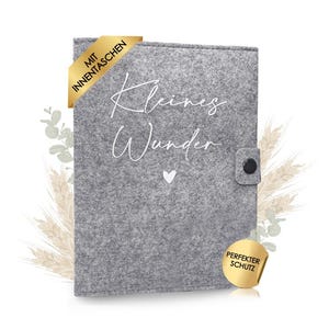 Könnte beinhalten: Eine graue Filzhülle mit dem weißen Schriftzug "Kleines Wunder" und einem kleinen Herzsymbol. Sie verfügt über einen schwarzen Knopfverschluss, ein goldenes Ecketikett mit "MIT INNENTASCHEN" und ein rundes goldenes Etikett "PERFEKTER SCHUTZ."