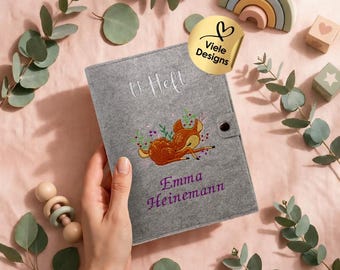 May include: A gray felt cover for a medical booklet featuring the embroidered text U Heft, a brown deer illustration, and the name Emma Heinemann in purple. The cover includes a snap button closure and a gold circular sticker.