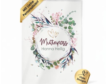 May include: A rectangular protective cover for a maternity record book featuring a floral wreath illustration in shades of green, pink, and blue. The text reads Mutterpass and Hanna Heilig, with two small heart outlines above the text.