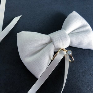 May include: A white satin bow tie with a gold ring and a clear stone. The bow tie is on a dark gray background.