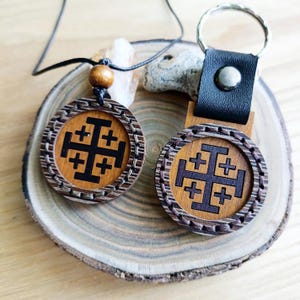 May include: A wooden pendant necklace and keychain featuring a Jerusalem cross design. The pendant hangs from a black cord with a wooden bead. The keychain has a black leather strap and metal ring. Both items have a brown wooden base with a decorative border.