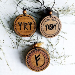 Puede incluir: Tres collares con colgantes de madera con cordones negros y cuentas de madera. Cada colgante tiene forma circular con símbolos y letras tallados. Los colgantes son marrones con diseños grabados en marrón oscuro o negro. El fondo incluye plantas secas.