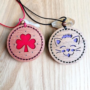 May include: Two wooden pendant necklaces. One has a red clover design, the other a cat face with purple accents. Both are round with stitched edges and hang on black and red cords.