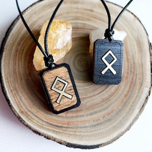 May include: Two rectangular wooden pendants on black cords. One pendant is light brown wood with a dark border, and the other is solid black wood. Both feature a metallic Othala rune symbol centered on the front face, suspended by a black cord.