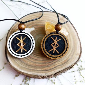 May include: Two circular wooden pendants on black cords. The left pendant has a white outer ring and a black center with a gold-toned engraved rune. The right pendant has a natural wood outer ring and a black center with a light brown engraved rune.