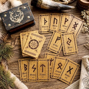 May include: A set of rectangular wooden cards featuring engraved Norse runes and English translations, accompanied by a dark wooden box with a wolf head design. The cards display symbols such as Thurisaz, Uruz, Wunjo, Fehu, Jera, Sowilo, Tiwaz, Othila, and Gebo.