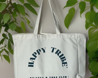 Puede incluir: Una bolsa de lona blanca con asas negras y el texto "HAPPY TRIBE" en un arco azul oscuro sobre el texto "PILATES & YOGA CLUB. EST 2014".