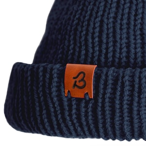 May include: A navy blue knitted beanie with a brown leather patch that has the letter "B" in a stylized font.