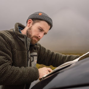 Könnte beinhalten: Ein Mann mit einer grauen Mütze und einer grünen Fleecejacke schaut auf eine Karte, während er neben einem schwarzen Auto steht.