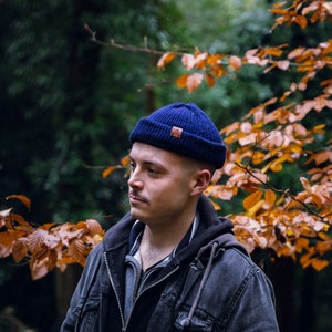 Peut inclure: Un homme portant un bonnet en laine bleu avec un patch en cuir marron sur le devant. Il porte une veste bleu foncé et se tient dans une forêt avec des feuilles d'automne.