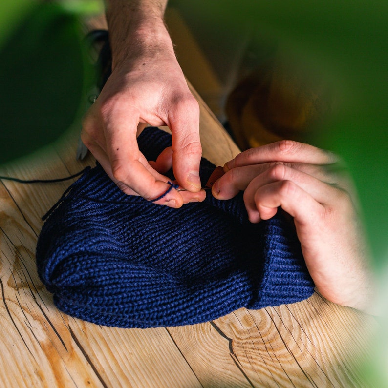 May include: A person is sewing a blue knitted beanie with a needle and thread. The beanie is on a wooden surface.