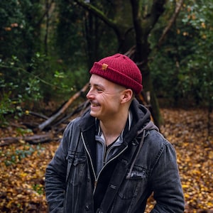 May include: A man wearing a red beanie and a dark denim jacket smiles while standing in a forest setting. The ground is covered in fallen leaves.