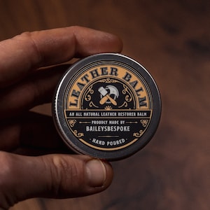 May include: A round, silver tin of leather balm held in a hand. The label features the words "LEATHER BALM" in a curved, gold font, with "AN ALL NATURAL LEATHER RESTORER BALM" below. The text "PROUDLY MADE BY BAILEYSBESPOKE HAND POURED" is also visible.