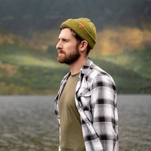 May include: A man wearing a green beanie and a black and white plaid shirt is standing in front of a body of water. The water is calm and reflects the sky.