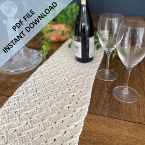 May include: A white macrame table runner with a diamond pattern, placed on a wooden table with two wine glasses and a bottle of wine.