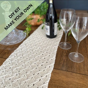 May include: A white macrame table runner with a diamond pattern. The runner is on a wooden table with two wine glasses and a bottle of wine.