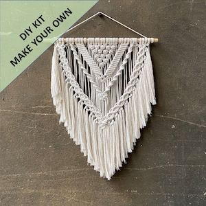 May include: DIY kit to make your own macrame wall hanging. The kit includes natural cotton cord and a wooden dowel. The finished wall hanging features a geometric pattern with fringe.