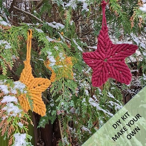 May include: DIY kit to make your own macrame star decorations. The kit includes two stars, one red and one yellow, hanging from a snowy evergreen tree. The text "DIY KIT MAKE YOUR OWN" is visible in the lower right corner of the image.