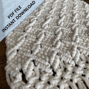 May include: A close-up of a white macrame wall hanging with a geometric pattern. The macrame is made with thick cotton cord and has a fringe at the bottom.