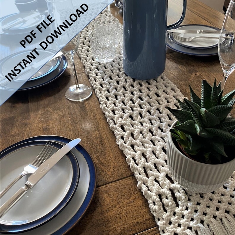 Pode incluir: Um corredor de mesa de croch&ecirc; branco com franjas em uma mesa de madeira. O corredor de mesa &eacute; disposto com um jarro azul, dois pratos brancos, dois copos, uma faca e um garfo de prata e uma pequena planta suculenta em vaso.