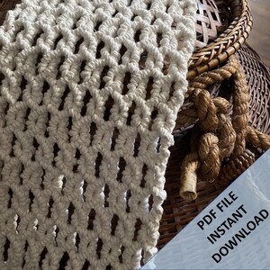 Pode incluir: Uma tape&ccedil;aria de macram&eacute; branca com um padr&atilde;o de diamante. O macram&eacute; est&aacute; pendurado sobre uma cesta tecida com uma corda.