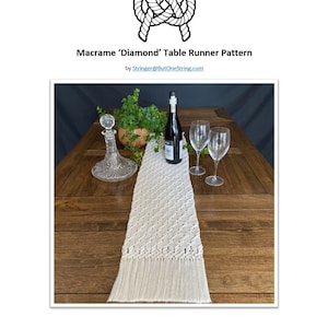 May include: A white macrame table runner with a diamond pattern and fringe. The runner is laid out on a wooden table with a glass decanter, wine bottle, and two wine glasses.