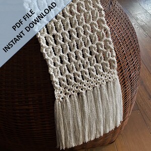 Pode incluir: Um corredor de mesa de macram&eacute; branco com um padr&atilde;o geom&eacute;trico e franjas. O corredor est&aacute; sobre uma superf&iacute;cie de vime marrom.