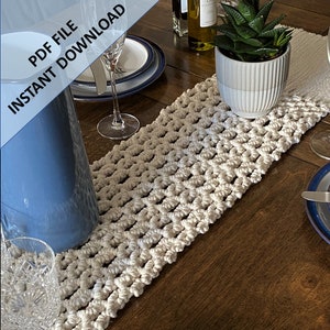 Pode incluir: Um corredor de mesa de croch&ecirc; branco com um padr&atilde;o geom&eacute;trico. O corredor est&aacute; sobre uma mesa de madeira com um jarro de vidro azul, um copo e uma suculenta em vaso.