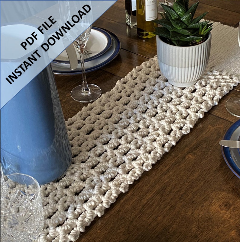 Pode incluir: Um corredor de mesa de croch&ecirc; branco com um padr&atilde;o geom&eacute;trico. O corredor est&aacute; sobre uma mesa de madeira com um jarro de vidro azul, um copo e uma suculenta em vaso.