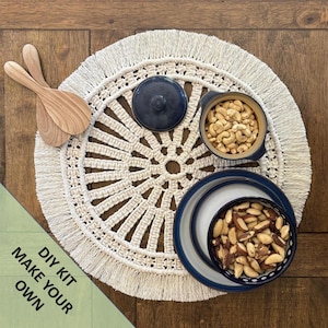 May include: A DIY kit to make your own macrame table mat. The kit includes a white macrame table mat with a fringe border, a blue bowl filled with cashews, a blue bowl filled with Brazil nuts, and two wooden spoons.