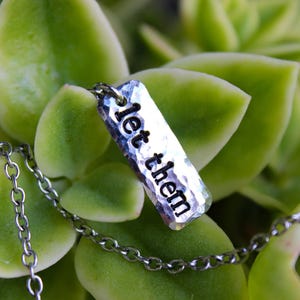 Puede incluir: Un collar plateado con un colgante rectangular que dice "let them" en negro. El colgante tiene una textura martillada y cuelga de una delicada cadena. El fondo es un grupo de hojas suculentas verdes.
