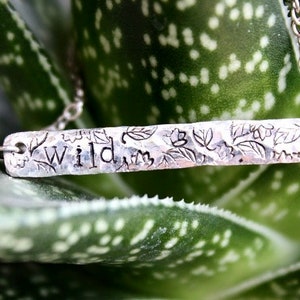 Könnte beinhalten: Silberne Halskette mit einem gehämmerten Barrenanhänger, auf dem "wild" mit floralen Designs steht. Die Halskette liegt auf einer grünen Pflanze.